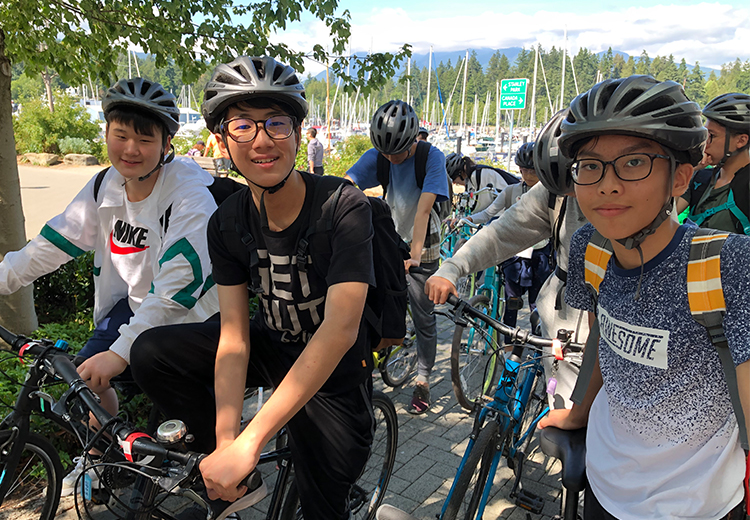 同學於溫哥華史丹利公園海邊騎單車，頭戴安全帽展現活力青春，背景可見遊艇與遠山景致 | 加拿大遊學團